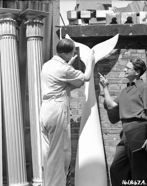 Two unidentified men are working on a mermaid tail, which will be used in the 1948 film "Miranda." There are columns and other building materials next to the tail, suggesting that this might be a prop or set-building shop. 