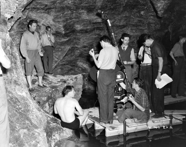 Actor Griffith Jones on the underwater cave set, preparing to film a scene from the 1948 movie "Miranda." The set contains a platform where some of the film crew is set up with a movie camera and a microphone above an artificial body of water.