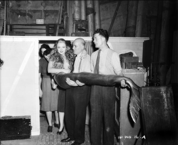 Glynis Johns wearing a mermaid costume is being carried by two unidentified male crew members. The location is probably a sound stage for the 1948 film "Miranda." Two other unidentified persons are standing behind her. 