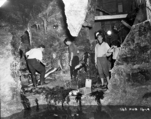 Actors Glynis Johns and Griffith Jones, and unidentified crew, on the underwater cave set of the 1948 film "Miranda." Johns is in her mermaid costume. The studio lights and sound stage are on the right. A picnic basket is in the foreground above the pool of water.