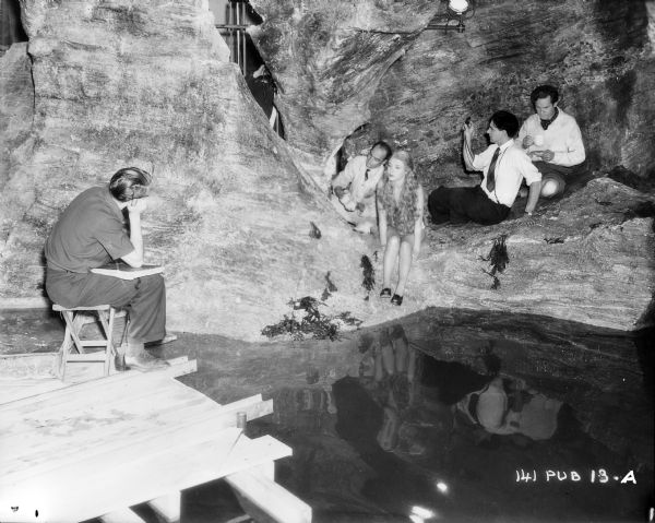 Glynis Johns, Griffith Jones, and the film crew on the underwater cave set of the 1948 film "Miranda." The sound stage and a light are set up above a pool of water. On the left is an unidentified man sitting on a platform built.