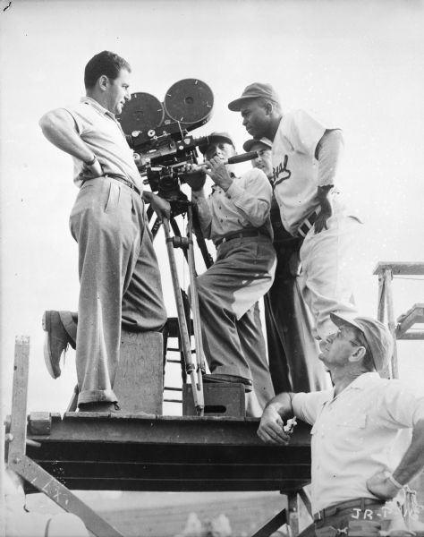 Jackie Robinson is looking at a movie camera on the set of the 1950 film "The Jackie Robinson Story." He and four other members of the film crew are standing on top of a raised platform, and another man standing on the ground is looking up at them.