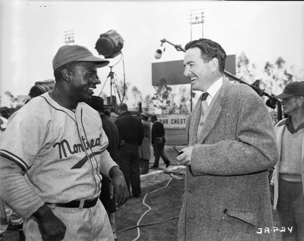 Jackie Robinson and an unknown man are standing on a baseball field during the making of the 1950 film "The Jackie Robinson Story." Behind them are men working with assorted pieces of film making equipment, including lights and a microphone. 