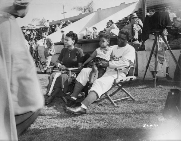 Jackie Robinson, his wife Rachel Robinson, and a child, probably their son Jackie Robinson Jr. They are sitting in chairs on a baseball field during the making of the 1950 film "The Jackie Robinson Story." Behind them are spectators and something, probably a camera, covered with black cloth on a tripod.