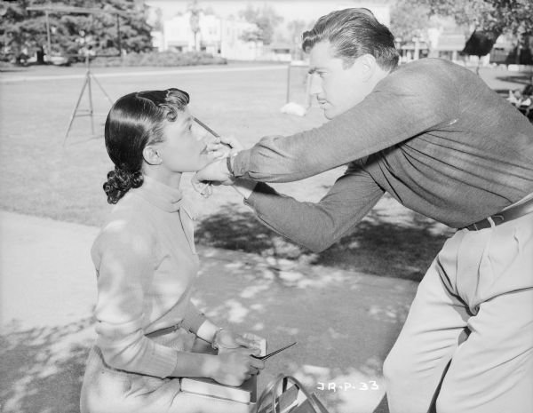Actress Ruby Dee, sitting outdoors, is having her makeup applied by an unidentified man, probably makeup artist Dave Grayson, during the making of the 1950 film "The Jackie Robinson Story." Behind them are assorted pieces of film making equipment. 