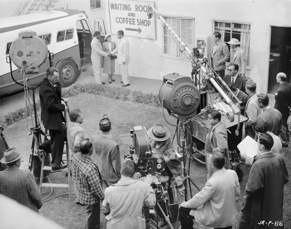 Elevated view of actors Jackie Robinson, Ruby Dee, and Roy Glenn filming a scene from the 1950 film "The Jackie Robinson Story." The trio are at a bus stop set, and are standing next to a bus. In the foreground is the assorted crew and film making equipment, including lights, a microphone, and a movie camera. 