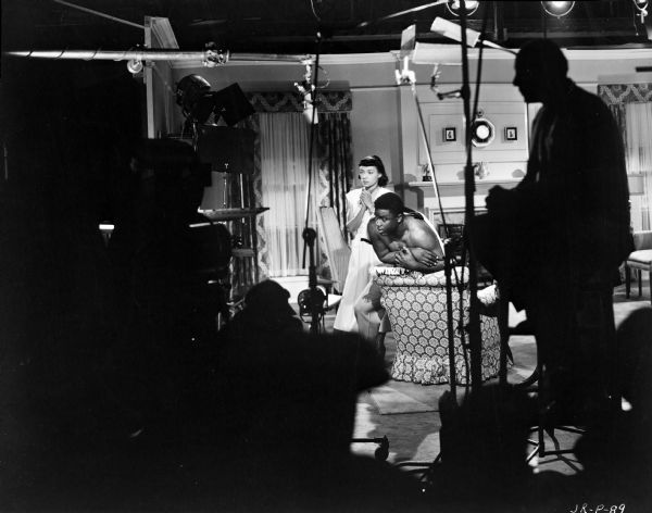 Actors Ruby Dee and Jackie Robinson pausing while filming a scene from the 1950 film "The Jackie Robinson Story." They are on a sound stage. In the foreground in silhouette are men working with film making equipment, including a microphone.