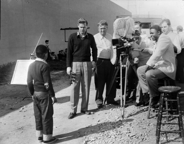Actors Howard Louis MacNeely and an unidentified man are filming a scene from the 1950 film "The Jackie Robinson Story." Jackie Robinson is sitting on a stool by a movie camera, which has its reels covered with cloth. The unidentified actor is holding a baseball glove.