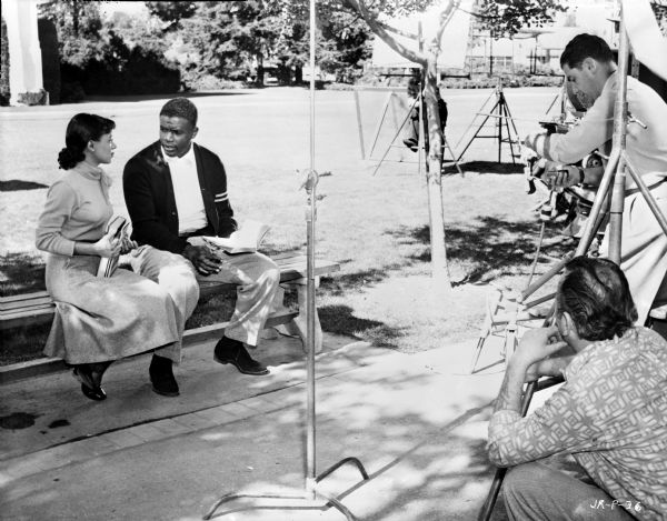 Actors Ruby Dee and Jackie Robinson filming a scene from the 1950 film "The Jackie Robinson Story." They are sitting on a bench outdoors. A reflector is on the grass behind them. Two men are behind equipment on the right.