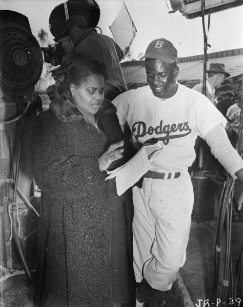 Actors Louise Beavers and Jackie Robinson are looking at a script together during the production of 1950 film "The Jackie Robinson Story." They are on a baseball field. Behind them a man is looking through a movie camera, and there are two film lights and people in the background on the right.