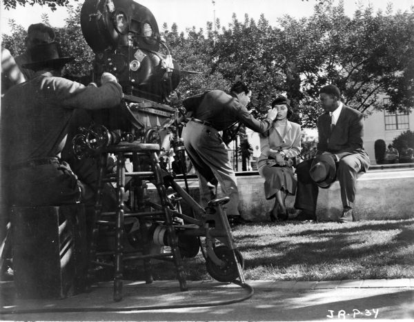 Actors Ruby Dee and Jackie Robinson outdoors filming a scene from "The Jackie Robinson Story." Dee is getting makeup applied or touched up, probably by makeup artist Dave Grayson. Men are standing behind a movie camera in the left foreground.