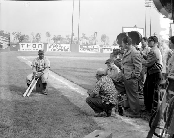 Jackie Robinson filming a scene from the 1950 movie "The Jackie Robinson Story." Robinson is sitting on a box on a baseball field holding two baseball bats. The film crew is on the right near a movie camera. 