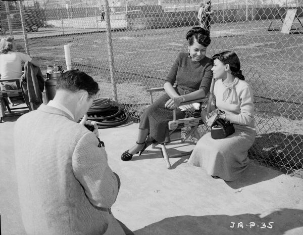 Rachel Robinson and actress Ruby Dee during the production of the 1950 film "The Jackie Robinson Story." They are in front of a fence at a baseball field. A photographer in the left foreground is taking pictures of them.