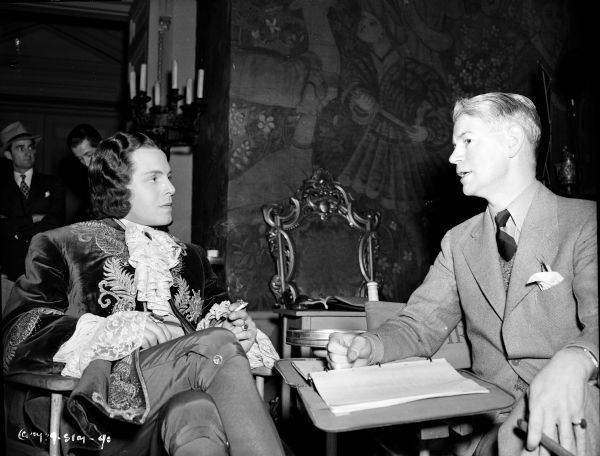 Actor Louis Hayward and director James Whale during the production of the 1939 film "The Man in the Iron Mask." Both men are sitting at a table on set, with what might be script. Behind them is a tapestry hanging from a set.