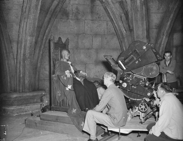 Director James Whale filming a scene from the 1939 film "The Man in the Iron Mask." An unidentified actor, possibly Louis Hayward, is wearing the iron mask and is sitting on a throne in a dungeon set. On the right, Whale and other crew members are next to a movie camera which is pointing towards the actor.