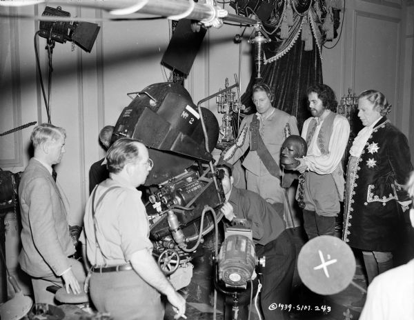 Actors Warren William, Louis Hayward, and Walter Kingsford in a scene from the 1939 film "The Man in the Iron Mask." Hawyard is holding the iron mask. The film crew and film making equipment are on the left, including lights, a microphone, and a movie camera.