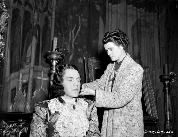 Actor Louis Hayward in costume having his hair prepared by an unidentified woman on the set of the 1939 film "The Man in the Iron Mask." Behind them are tapestries and candlesticks.