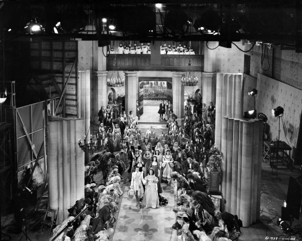 Elevated view of the filming of the wedding scene from the 1939 film "The Man in the Iron Mask." Actor and extras are gathered on a large, two level church interior set. The sound stage and assorted film equipment (including lights and a microphone) are on the far left, far right and top.