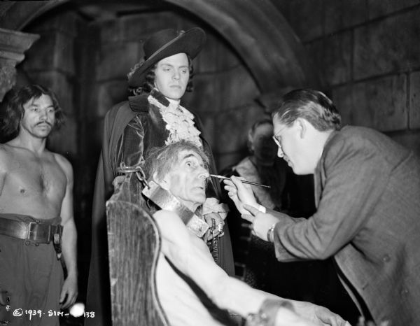 Actor Louis Hayward watching an unidentified makeup artist working on actor D'arcy Corrigan on a prison set for the 1939 film "The Man in Iron Mask."