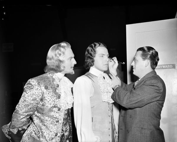 Actor Louis Hayward having makeup applied to his face by an unidentified man, next to actor Joseph Shildkraut. Both are in costume for the 1939 film "The Man in the Iron Mask." Hayward's name is on a door behind them, indicated that they are probably outside his dressing room.