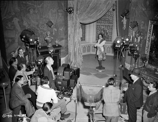 Elevated view of actor Louis Hayward during the making of the 1939 film "The Man in the Iron Mask." He is standing on a short stool, and there are tapestries and a window behind him. The film crew is in the foreground surrounding Hayward, along with lights and a movie camera pointing towards him.