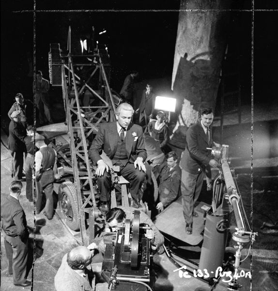 Director and actor Laurence Olivier sitting on a camera crane on the set of the 1949 film "Hamlet." On the right is a man working a boom microphone attached to a device, possibly a boom pole on a stand with wheels.