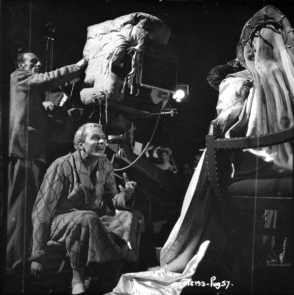 Director and actor Laurence Olivier crouching under a movie camera. He is directing an actress, probably Eileen Herlie, whose face is obscured. The camera is wrapped in fabric.