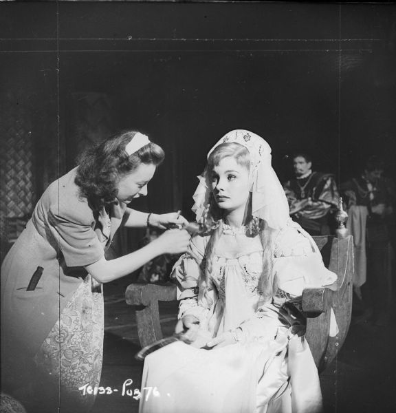 Actress Jean Simmons getting her hair adjusted by an unidentified woman, probably hairdresser Vivienne Walker. Simmons is sitting in costume on the set of the 1948 movie "Hamlet."
