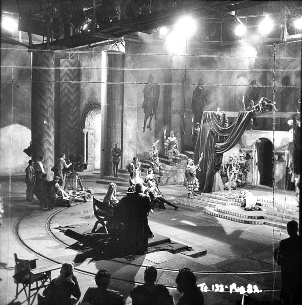 Elevated view of the cast and crew filming the play scene from the 1949 film "Hamlet." The movie camera is on a semicircular track around the stage. Lights are along the ceiling.