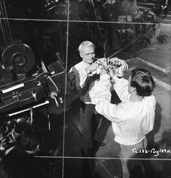 Elevated view of actors Laurence Olivier and Terence Morgan filming the duel scene from the 1949 film "Hamlet." Both men are using daggers and rappers. A movie camera is on the left. A group of people are watching in the background.