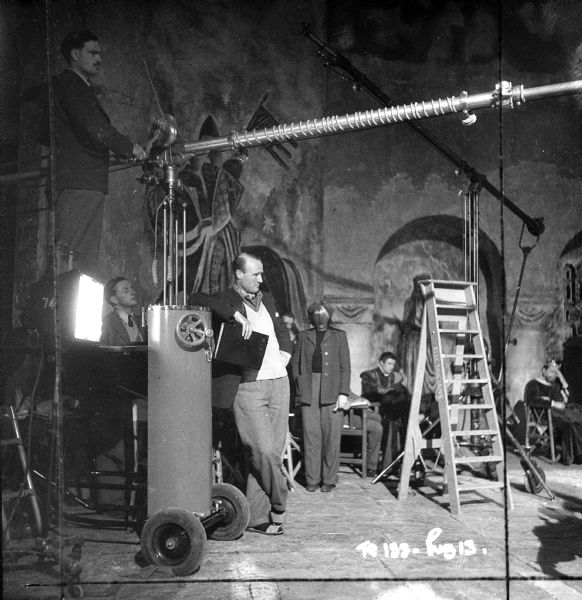Recording equipment, most likely a microphone boom pole attached to a stand with wheels, on the set of the 1949 film "Hamlet." Assorted cast and crew are scattered around the room. 