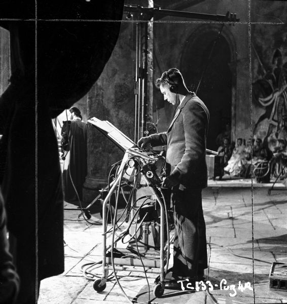 An unidentified person is standing and working with what is probably sound recording equipment on the set of the 1949 film "Hamlet." A boom mic is behind him. Actors and actresses are in the background.