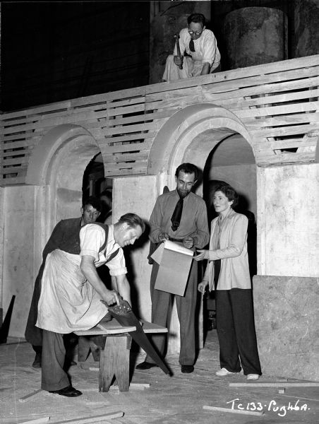 Art Director Carmen Dillon and an unidentified man are viewing the construction of one of the sets from the 1948 film "Hamlet." Another unidentified man is hammering on top of partially constructed arches, while another is sawing wood.