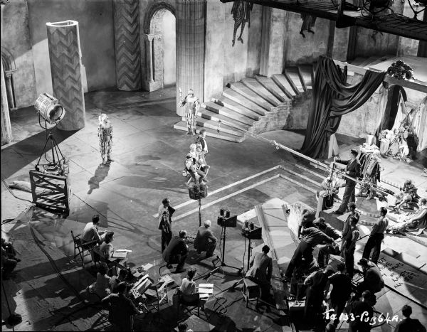 Elevated view of cast and crew filming a scene from the 1949 film "Hamlet." Laurence Olivier and Norman Wooland have a microphone over their heads. A stage for the play scene is on the right. A movie camera is covered with fabric and on a track at the bottom right.
