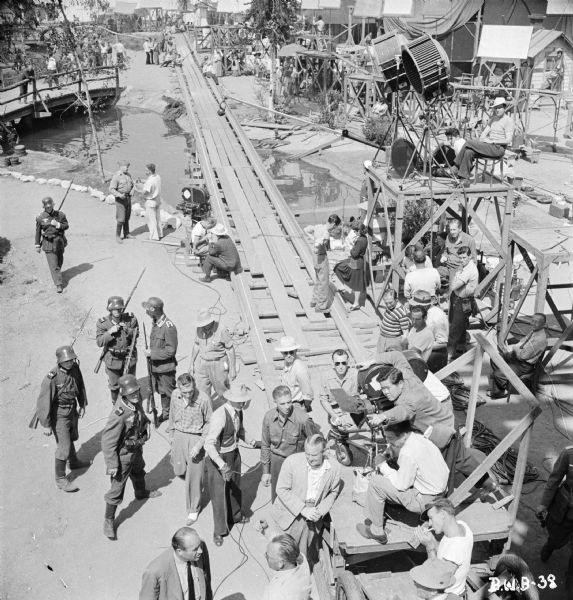 Elevated view of assorted cast and crew on a village set of the 1943 film "The North Star." Cinematographer James Wong Howe, in the lower right corner near a movie camera, is on a camera dolly. The dolly is on a long track that goes over an artificial lake. Another movie camera is further down the track. There are also lights and numerous reflectors on platforms.