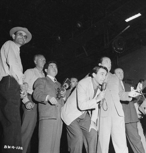 A group of men on a sound stage, watching something out of frame, probably the filming of the 1943 film "The North Star." Director Lewis Milestone is to the right of an unidentified man in the center holding a microphone. Cinematographer James Wong Howe is to the left of this man, and is holding an unidentified camera-like device, possibly a viewfinder.