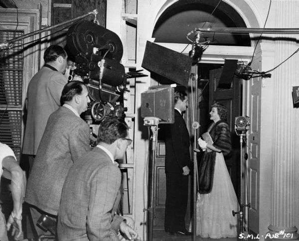 Actors Robert Cummings and Claudette Colbert filming a scene from the 1948 film "Sleep, My Love." They are surrounded by assorted film making equipment, including at least one microphone and lights. On the left are men standing near a movie camera.
