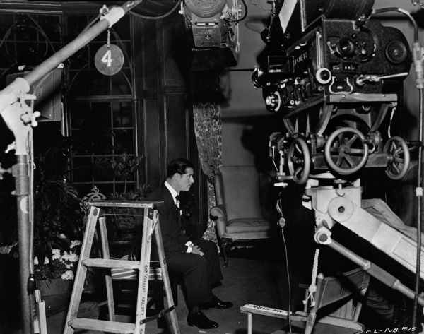 Actor Don Ameche sitting on the set of the 1948 film "Sleep, My Love." A light and movie camera are in the foreground. 