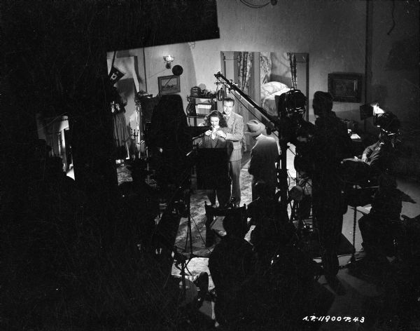 Elevated view of Dick Powell and Linda Darnell on the set of the 1944 film "It Happened Tomorrow." In silhouette in the foreground are the film crew and assorted equipment, including lights and a microphone over Powell and Darnell.