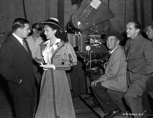 Actress Linda Darnell and director Réne Clair (far left) during the production of the 1944 film "It Happened Tomorrow." Men are near a movie camera in the center.