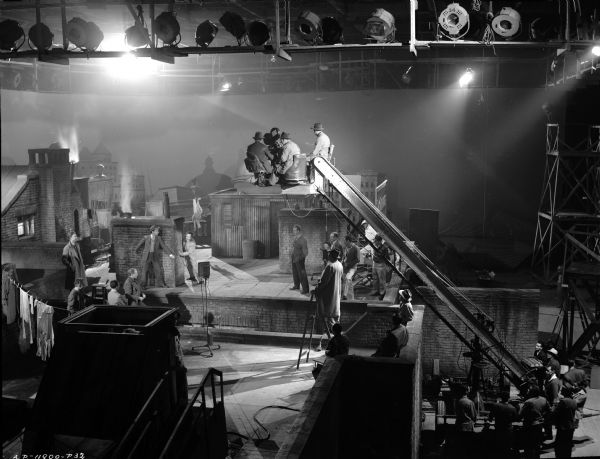 Elevated view of cast and crew filming a scene from the 1944 film "It Happened Tomorrow." The set recreates apartment rooftops. Studio lights are above the crew, and three people are working a movie camera on a platform at the top of a crane. 