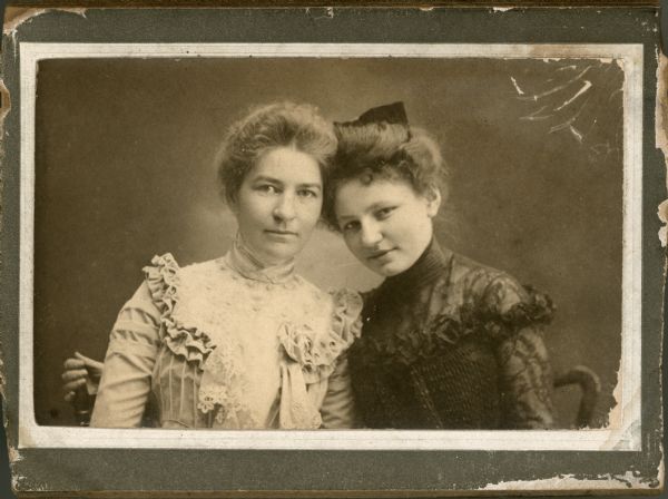 A head and shoulders portrait of Emma Brumder (1870-1962), left, and Amanda Zimmerman, right, later Mrs. Richard Rockwood. The women are in an affectionate pose, with their heads resting together, and are wearing fancy blouses trimmed with lace and frills. Amanda has a bow in her hair.  