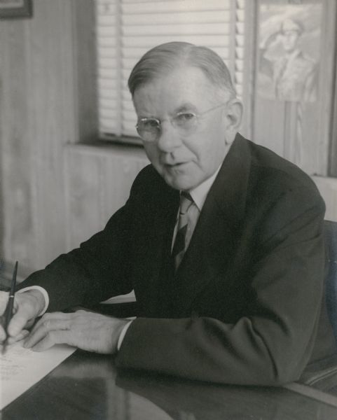 A quarter-length portrait of Herman Otto Brumder (1880-1950) seated at his desk, posed as if signing a document. There is a military poster on the wall behind him. He is wearing a suit and tie. A son of publisher George Brumder (1839-1910), H.O. Brumder was president of the Pressed Steel Tank Company of West Alllis.