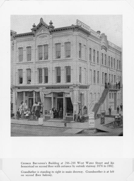 A halftone print copy of a photograph of George Brumder's 1873 three story brick building at 286-288 West Water Street. Brumder and his family lived on the second floor. As noted in the caption, George Brumder is pictured at right in the doorway at ground level. His wife, Henriette, is standing at left at the top of the stairway on the side of the building. Two signboards are standing on the wooden sidewalk near the corner. A sign along the top of the building reads: Germania Commercial, Book, and Job Printing. There is a small carriage at lower right. Brumder built a second building, known as the Brumder Block, in 1892, and the larger Germania Building in 1896 to house his growing German-language publishing business.