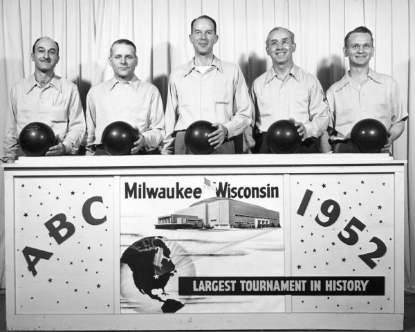 Five men holding bowling balls stand behind a sign identifying the 1952 A. B. C. [American Bowling Congress] Tournament. The man standing second from right is wearing a shirt with a bowling pin and the initials ABC embroidered on the sleeve. In the center of the sign is a drawing of the Milwaukee Arena, now the UW Milwaukee Panther Arena, which opened in 1950. There is also a drawing of a globe with a star identifying Milwaukee, and a line of print declaring "Largest Tournament in History." On the reverse is written "L to R  Kujawa, Patten, Brumder, Wittman, Sloan." Brumder was Edward J. Brumder (1907-1998), at that time vice president of The North American Press of Milwaukee.