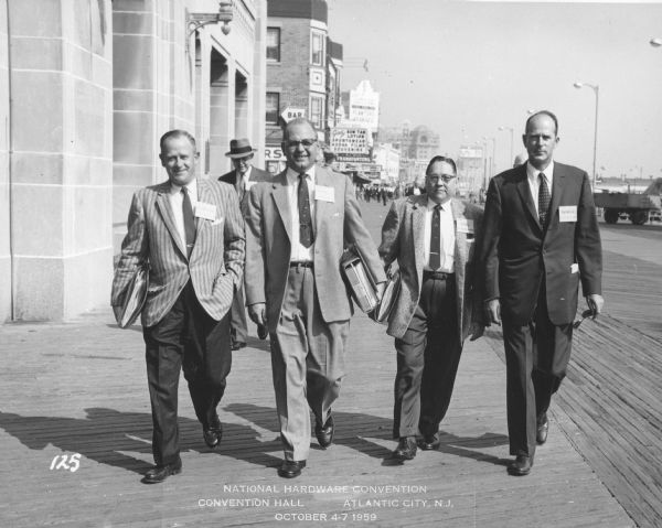 Five men in coats and ties are walking on the boardwalk. All of the men are wearing name badges, and three are carrying binders. A caption at the bottom reads: "National Hardware Convention, Convention Hall, Atlantic City, N.J., October 4-7 1959." At far right is Edward J. Brumder of Milwaukee, who at the time of the photograph was vice-president of The North American Press, which specialized in printing hardware catalogs. In the background are signs for a theater, a bar, and Planter's Peanuts.