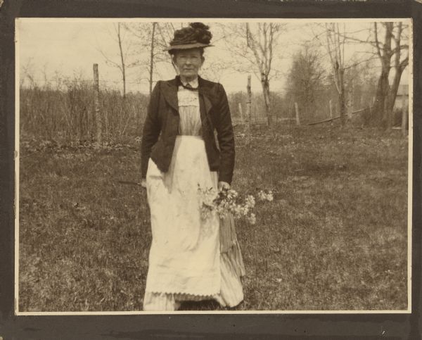 Henriette Brandhorst (Mrs. George) Brumder posing outdoors with a bouquet of blossoms in her left hand. She is wearing a short fitted jacket over a striped dress and apron. She is also wearing a fancy hat. There is a wire fence in the background. 
