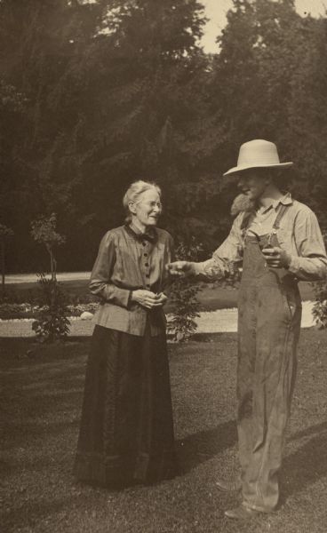 John Merker (1900-1961), dressed in straw hat, bib overalls and a work shirt, is holding a pipe in his left hand. He is wearing a fake beard. His grandmother, Henriette Brandhorst (Mrs. George) Brumder, appears to be amused.  