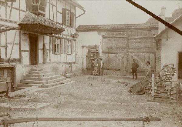 Several men and children are standing in an arched doorway at the rear of a small courtyard. On the left, steps lead to the door of a <i>Fachwerk</i> (timberframe) house. Two unidentified well-dressed men are standing on the right. There is a pile of bricks and a grindstone on the right. The house may have been owned by a relative of Milwaukee publisher George Brumder. Brumder, with his wife Henriette and two of their children, traveled from Milwaukee to his hometown of Breuschwickersheim in 1889. At that time a part of the German Empire, Breuschwickersheim is in the department of Bas-Rhin, region of Alsace, France.  
