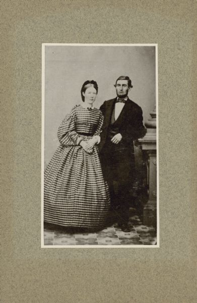 Full-length studio portrait of George Brumder and his wife, Henriette Brandhorst Brumder, taken around the time of their wedding, July 16, 1864. She is wearing a checked dress with a full skirt and full sleeves. He is wearing a suit and vest, and rests his left elbow on a decorative plinth. His right hand is on his wife's shoulder.  